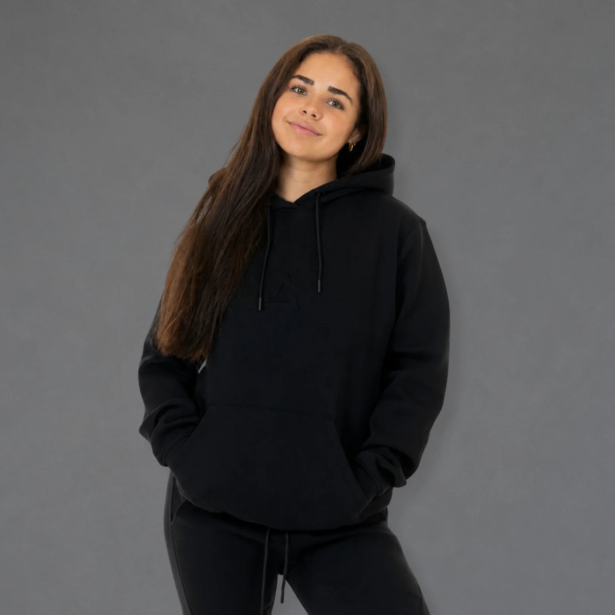 A woman with long brown hair wearing a black pullover hoodie and matching black sweatpants, standing against a solid gray studio background.