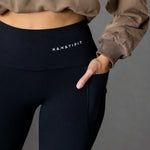 Close-up of a person wearing black high-waisted leggings with a white KANATIFIT logo on the waistband, paired with a brown sweatshirt, against a solid gray background.