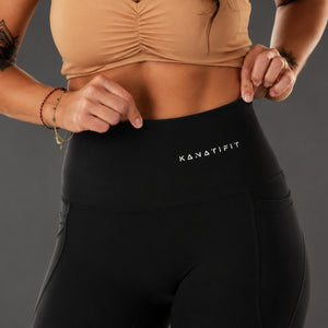 Close-up of a model wearing black high-waisted leggings with the white 'KANATIFIT' logo on the front, paired with a beige ruched sports bra against a dark gray studio background.
