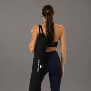 A woman shown from behind wearing a black sports bra and dark leggings while carrying a long black resistance band over her shoulder against a plain gray studio background.