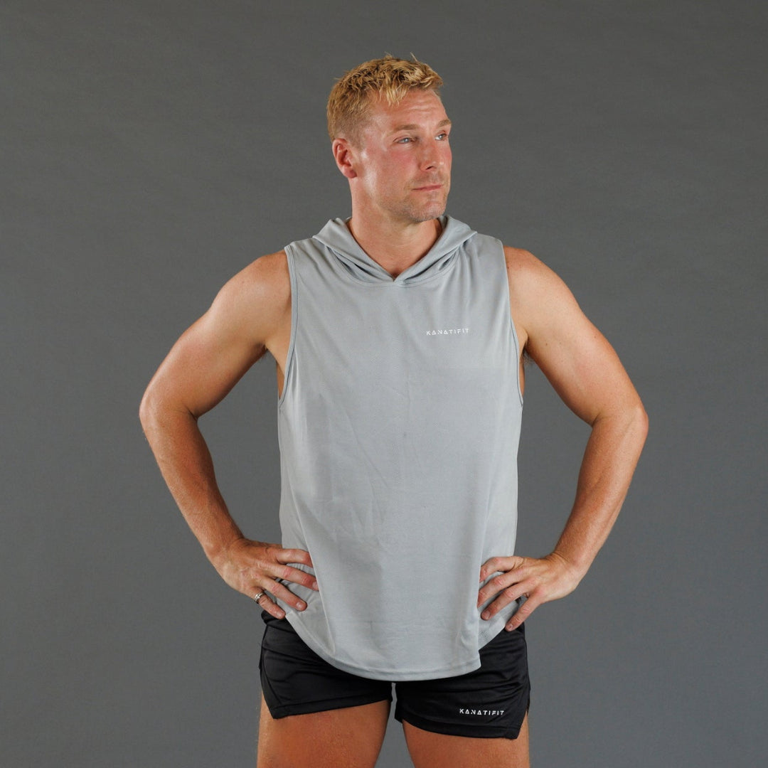 A man wearing a light gray sleeveless hooded athletic tank top and black athletic shorts, standing with hands on hips against a solid gray studio background.