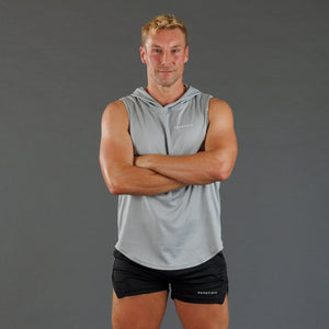A man wearing a light gray sleeveless hooded athletic top and black shorts, standing with arms crossed against a solid gray studio background. The brand name 'KANATIFIT' is visible on the chest and shorts.