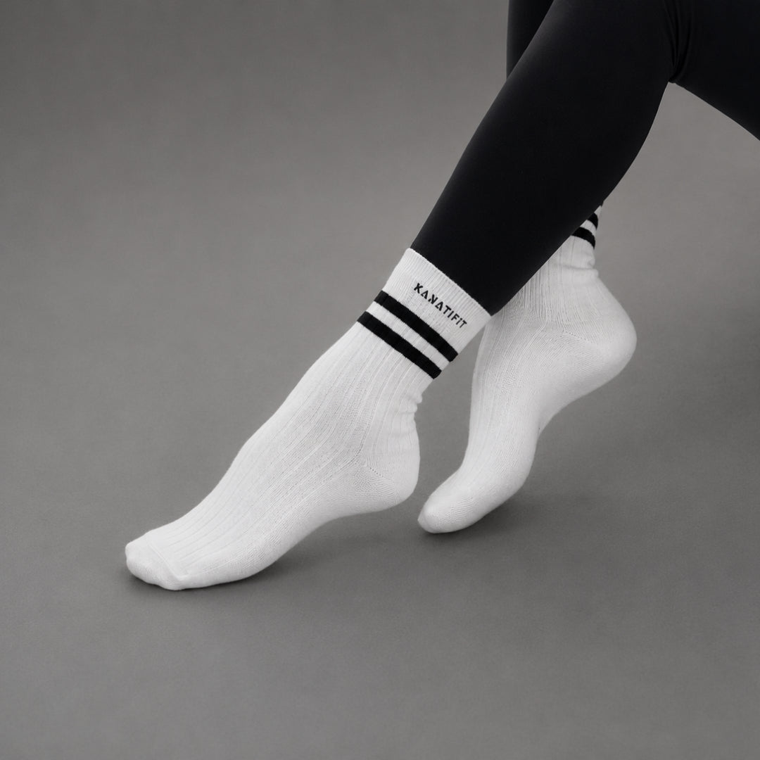 White ankle socks with black stripes and the brand name 'KANATIFIT' printed on the cuff, worn with black leggings against a neutral gray background.