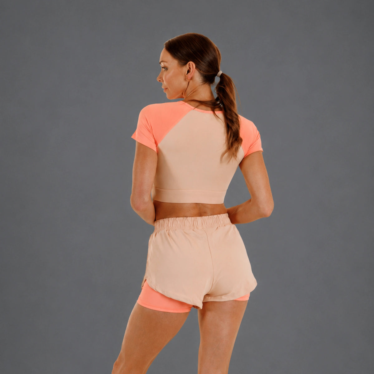 A woman shown from the back wearing a peach and beige athletic crop top and matching shorts with a coral inner layer, set against a solid gray studio background.