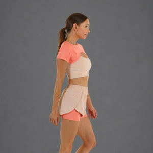 A woman in side profile wearing a coral and cream athletic set, consisting of a short-sleeve crop top with a chest cutout and matching shorts, against a solid gray studio background.