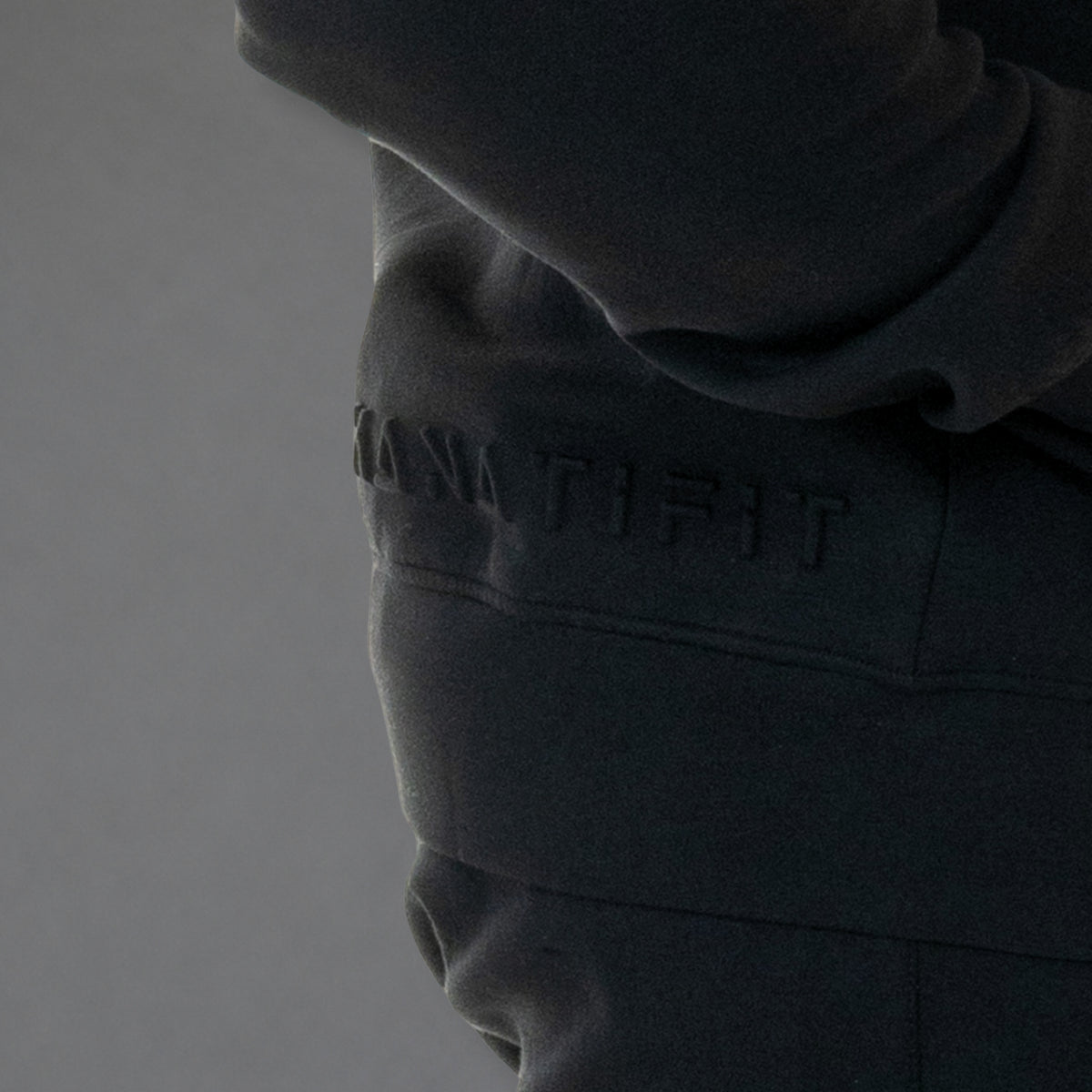 Close-up of a dark charcoal gray hoodie with the brand name 'KANATIFIT' embossed on the chest, shown on a model against a neutral gray background.