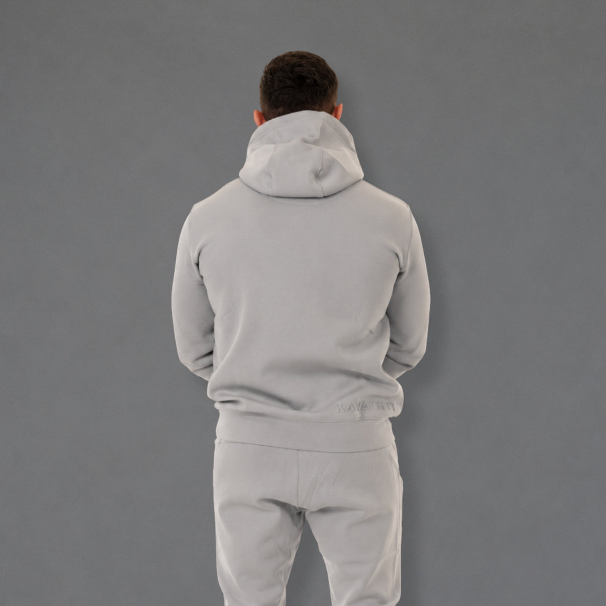 A man viewed from behind wearing a light gray hoodie and matching sweatpants against a solid gray studio background.
