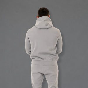 A man viewed from behind wearing a light gray hoodie and matching sweatpants against a solid gray studio background.
