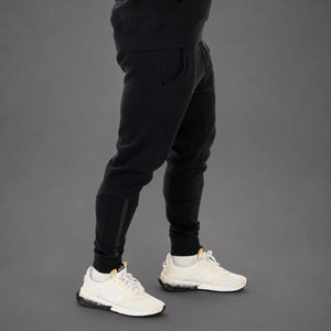 A person wearing black jogger-style pants with side compression panels and white sneakers, shown from the waist down against a dark gray studio background.