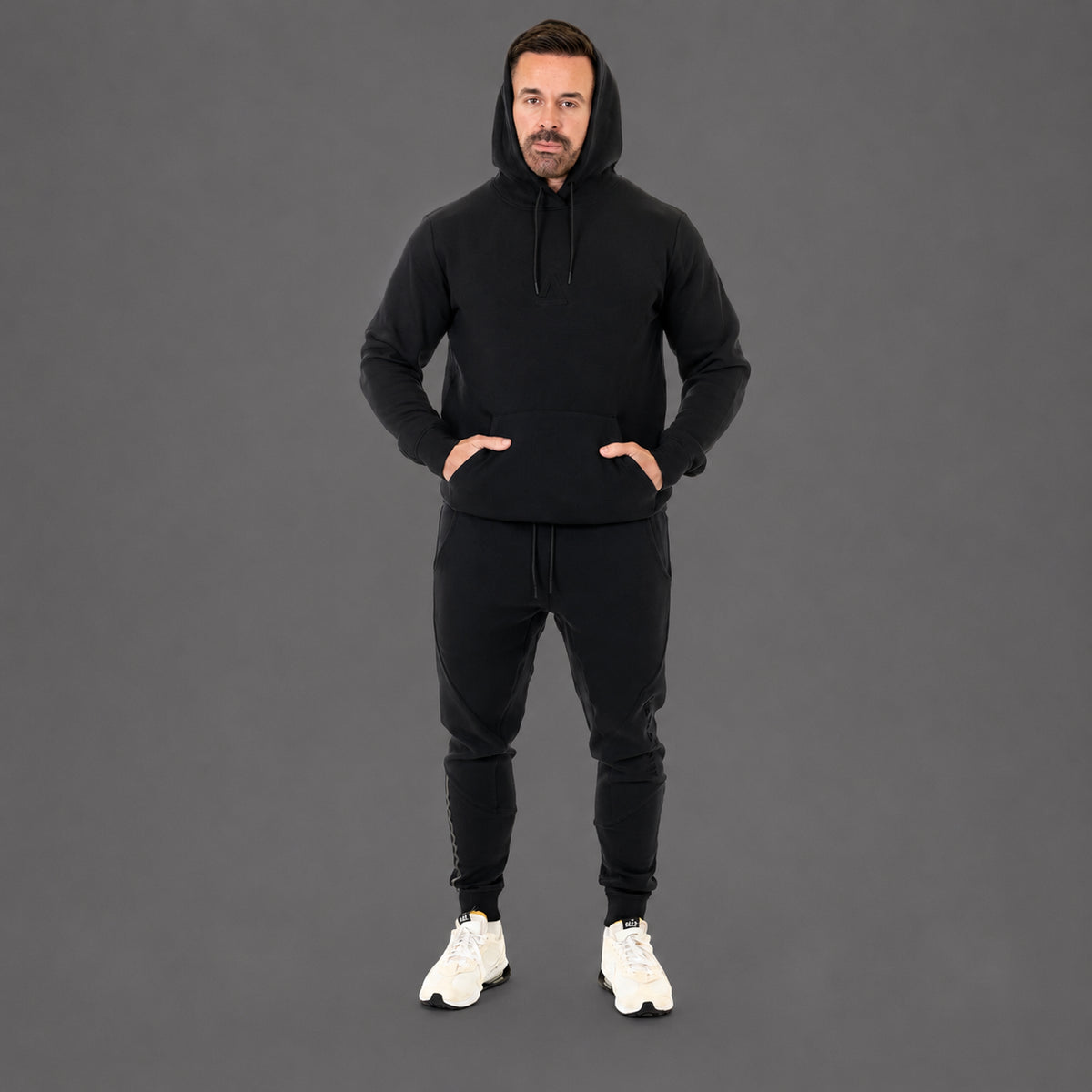 A man wearing a black pullover hoodie and matching black jogger pants, standing against a solid dark gray studio background. He is wearing white sneakers.