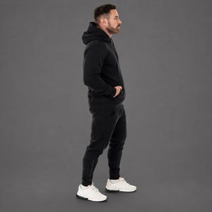 A man in a black hooded sweatshirt and black jogger pants stands in profile against a solid gray studio background, wearing white sneakers.