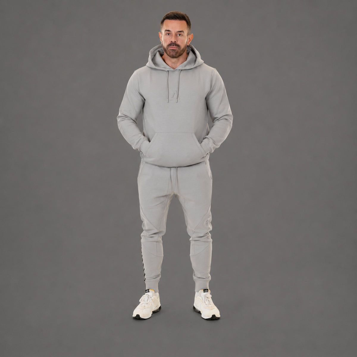 A man modeling a light gray two-piece tracksuit consisting of a pullover hoodie and matching jogger pants, paired with white sneakers, against a solid gray studio background.