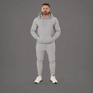A man modeling a light gray two-piece tracksuit consisting of a pullover hoodie and matching jogger pants, paired with white sneakers, against a solid gray studio background.