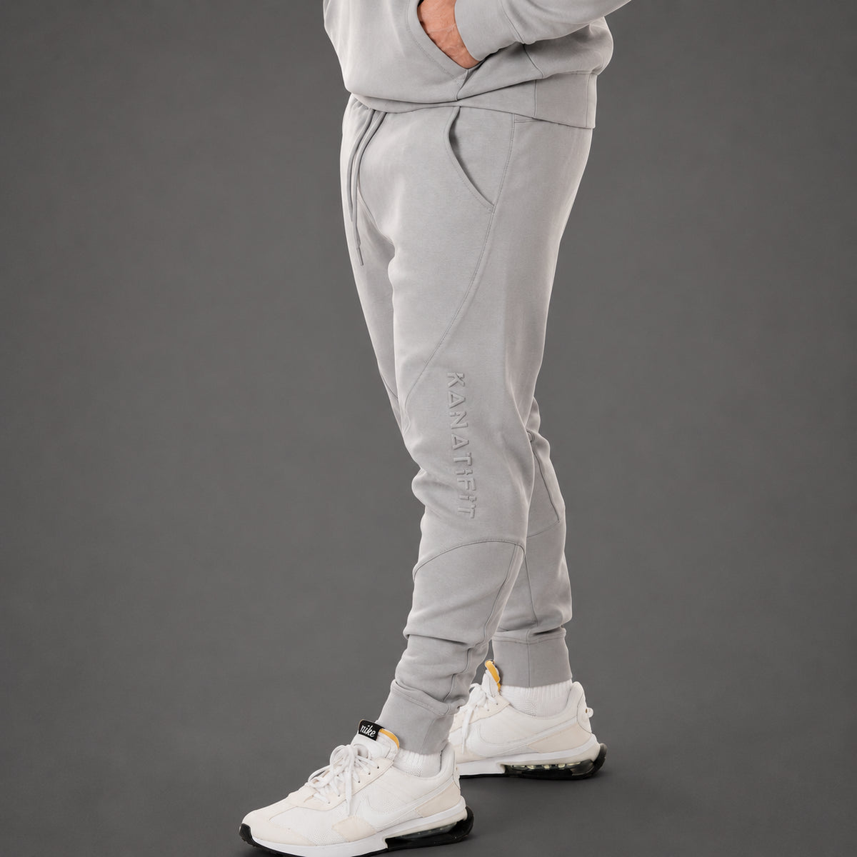A person wearing light gray jogger sweatpants with a vertical 'KANATIFIT' logo on the left leg, paired with white sneakers, shown from the waist down against a dark gray studio background.