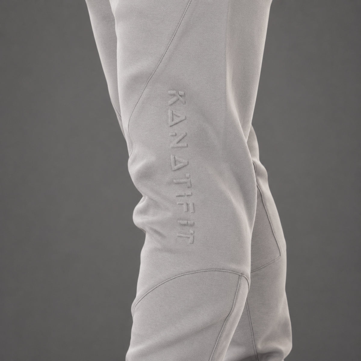 Close-up of light gray athletic pants worn on a model's leg, featuring an embossed vertical 'KANATIFIT' logo on the side, against a solid dark gray background.