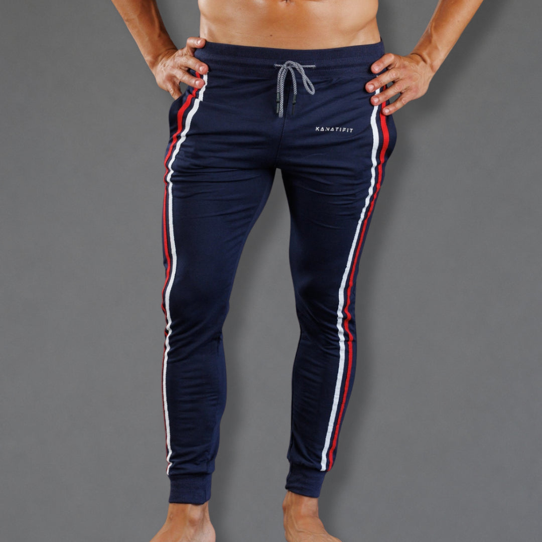 A man wearing navy blue track pants with red and white side stripes, standing against a solid gray background.