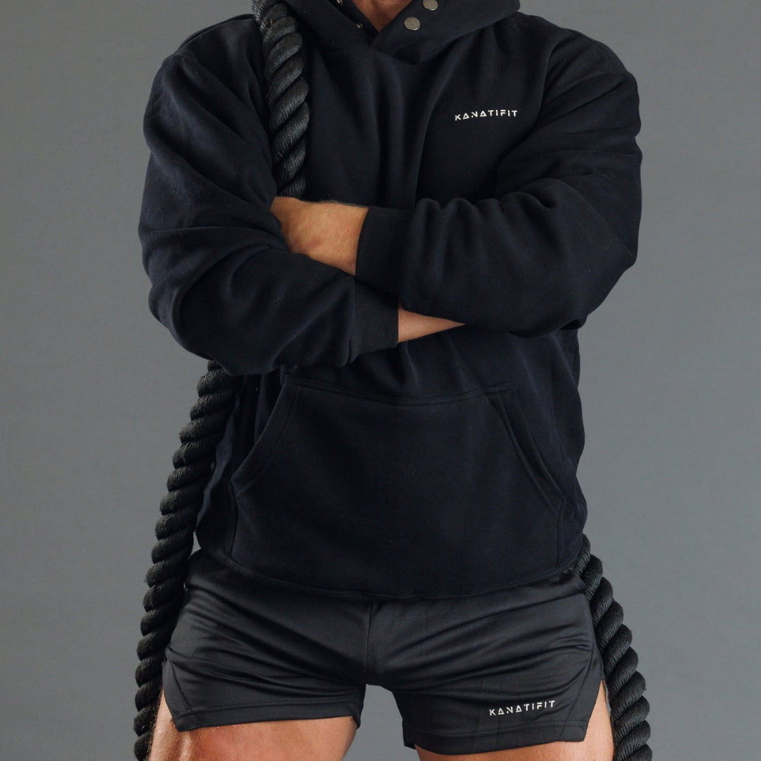 A person wearing a black KANATIFIT pullover hoodie and matching black athletic shorts, standing with arms crossed while holding a thick black battle rope against a plain gray studio background.