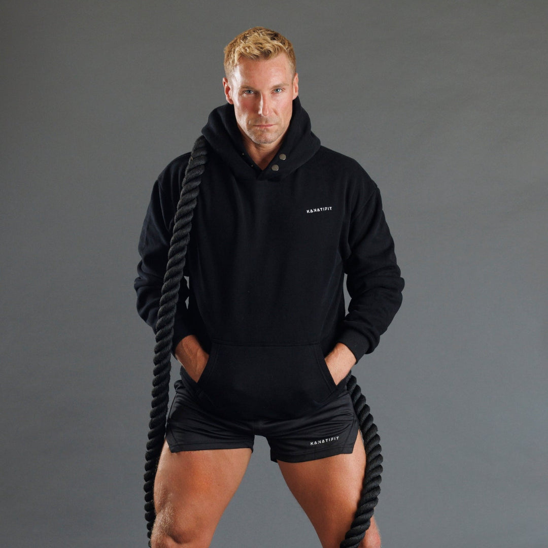 A man wearing a black athletic hoodie and matching black shorts stands against a dark gray studio background, holding a thick black battle rope. The hoodie and shorts have small white KANATIFIT branding on the chest and leg.