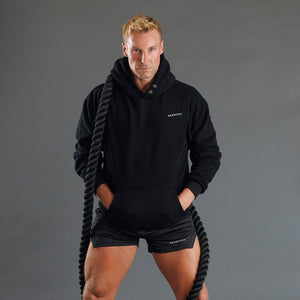 A man wearing a black athletic hoodie and matching black shorts stands against a dark gray studio background, holding a thick black battle rope. The hoodie and shorts have small white KANATIFIT branding on the chest and leg.