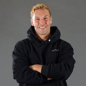 A smiling man with blond hair wearing a black pullover hoodie with a small white 'KANATIFIT' logo on the left chest, posed with arms crossed against a solid gray studio background.