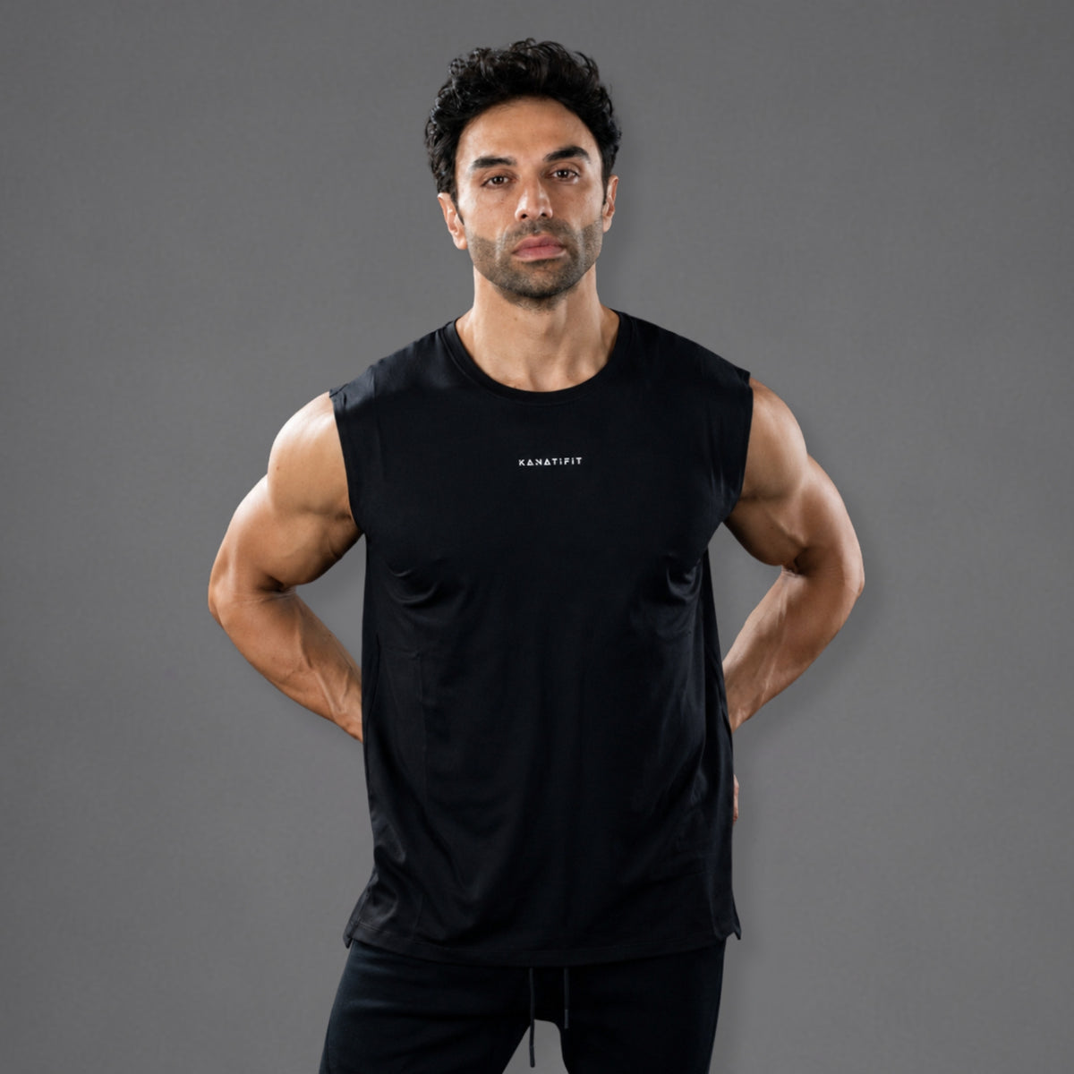 A muscular man wearing a black sleeveless athletic tank top with a small white 'KANATIFIT' logo on the chest, posed against a solid dark gray studio background.
