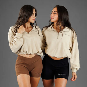 Two women modeling matching beige quarter-zip pullovers and fitted shorts against a plain gray studio background. One model wears brown shorts and the other wears black shorts, both with small white 'KANATIFIT' branding visible on the chest and shorts.