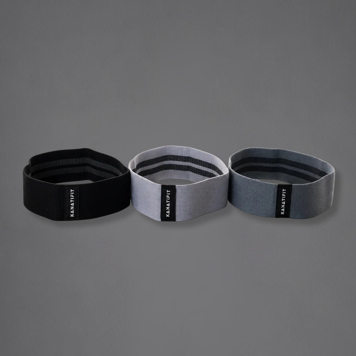 Three resistance bands in black, light gray, and dark gray, arranged side-by-side on a solid gray background. Each band features a black vertical label with the brand name 'KANATIFIT' in white.