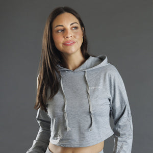A woman wearing a grey cropped hoodie with long sleeves and drawstrings, photographed against a solid gray studio background.
