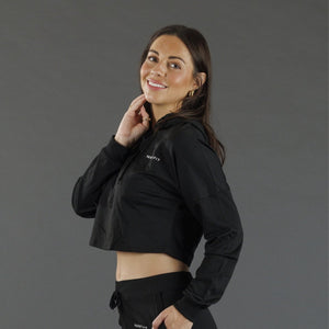 A woman modeling a black cropped zip-up hoodie with a small white 'KANATIFIT' logo on the chest, paired with black athletic pants, against a solid dark gray studio background.