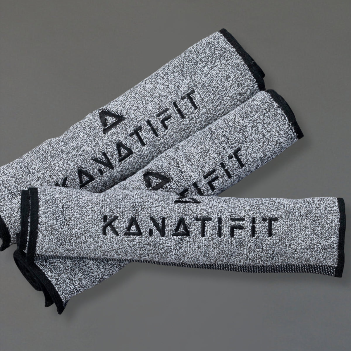 Three gray heathered sweat towels with black trim and the brand name KANATIFIT printed in black, arranged diagonally on a solid gray background.