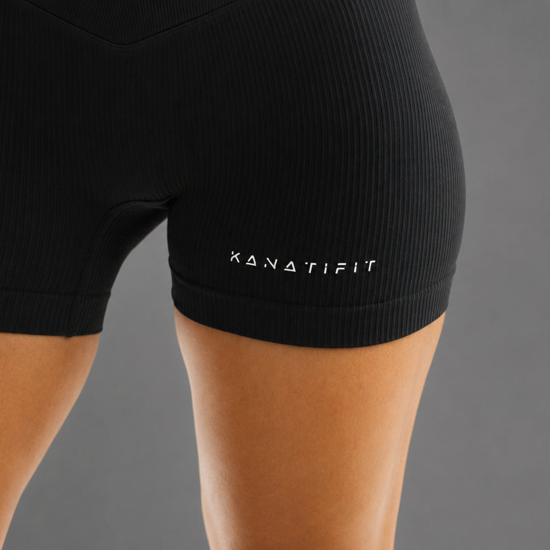 Close-up of a person wearing black ribbed athletic shorts with the brand name KANATIFIT printed in white on the side, against a plain gray background.
