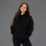A woman with long brown hair wearing a black pullover hoodie and matching black sweatpants, standing against a solid gray studio background.
