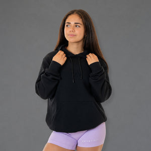 A woman wearing a black pullover hoodie with drawstrings and a front kangaroo pocket, paired with light purple fitted shorts, standing against a solid gray studio background.
