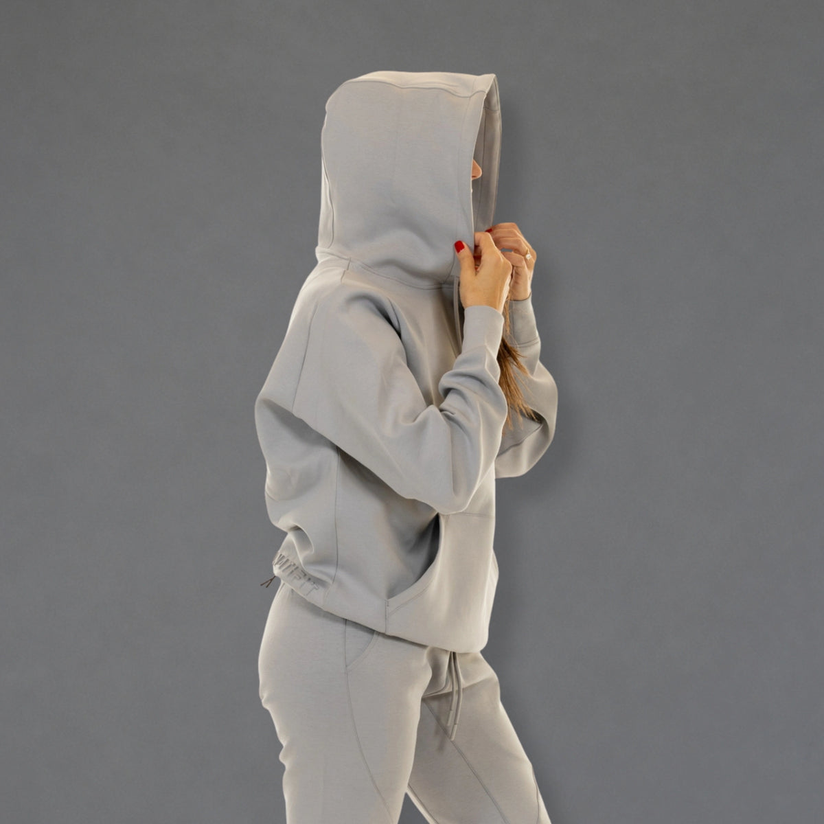 A person wearing a light gray hooded sweatshirt and matching jogger pants, shown from the side against a plain gray studio background.