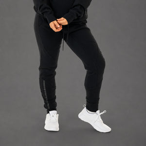 Black jogger sweatpants worn by a model, paired with a black hoodie and white sneakers, shown from the waist down against a solid gray studio background.