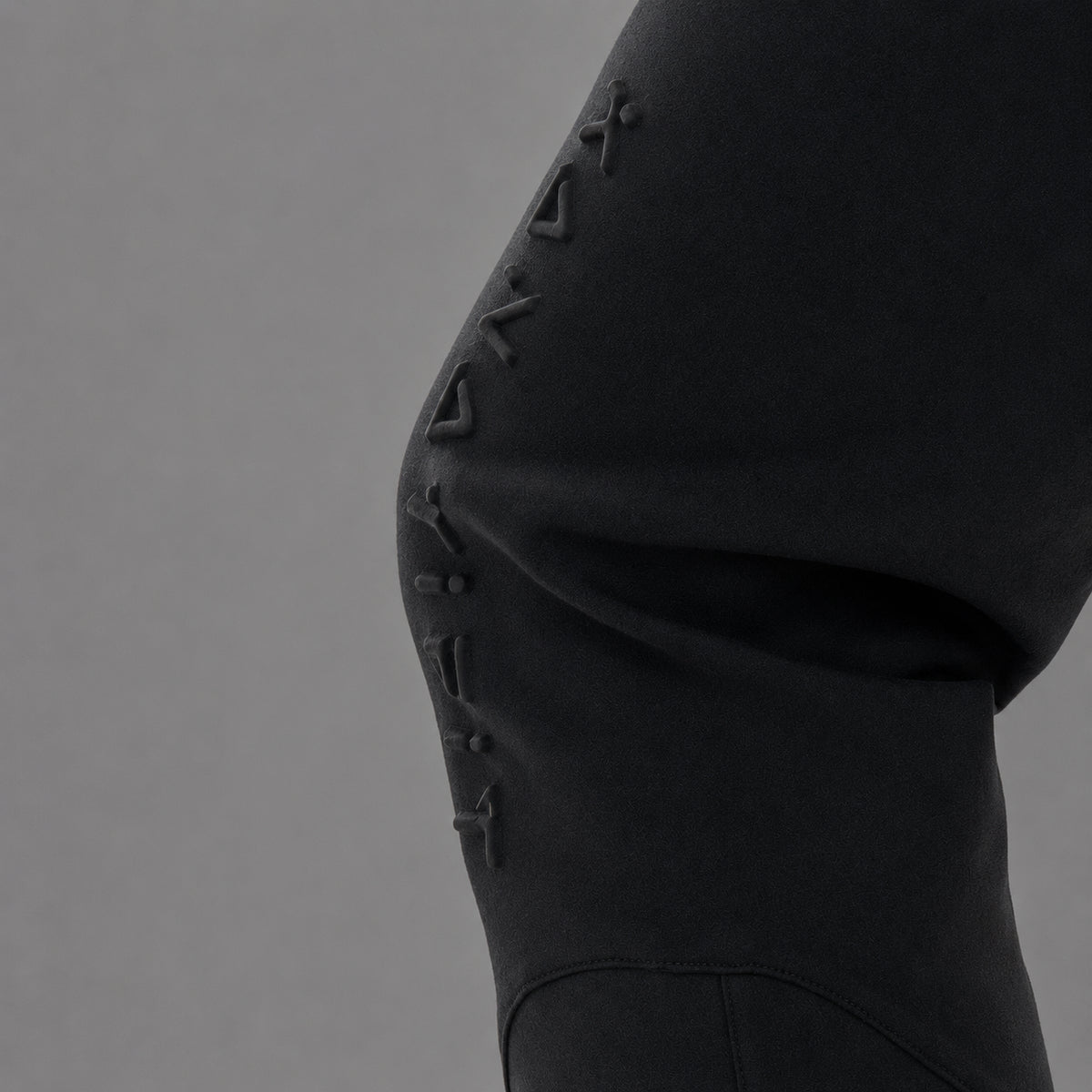Close-up of a black athletic garment, likely leggings or a skirt, showing a curved seam with raised geometric logo details running vertically along the side against a plain gray background.