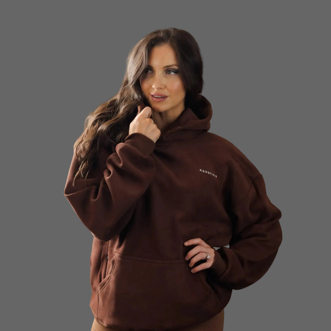 A woman modeling a dark brown fleece hoodie with a small white embroidered logo on the left chest, set against a solid dark gray studio background.