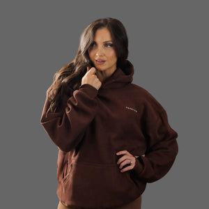 A woman modeling a dark brown fleece hoodie with a small white embroidered logo on the left chest, set against a solid dark gray studio background.