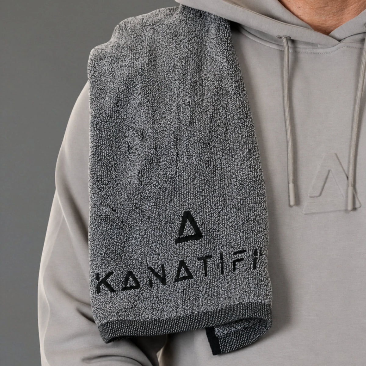 A gray heathered towel with black 'KANATIFIT' branding draped over the shoulder of a person wearing a light gray hoodie.