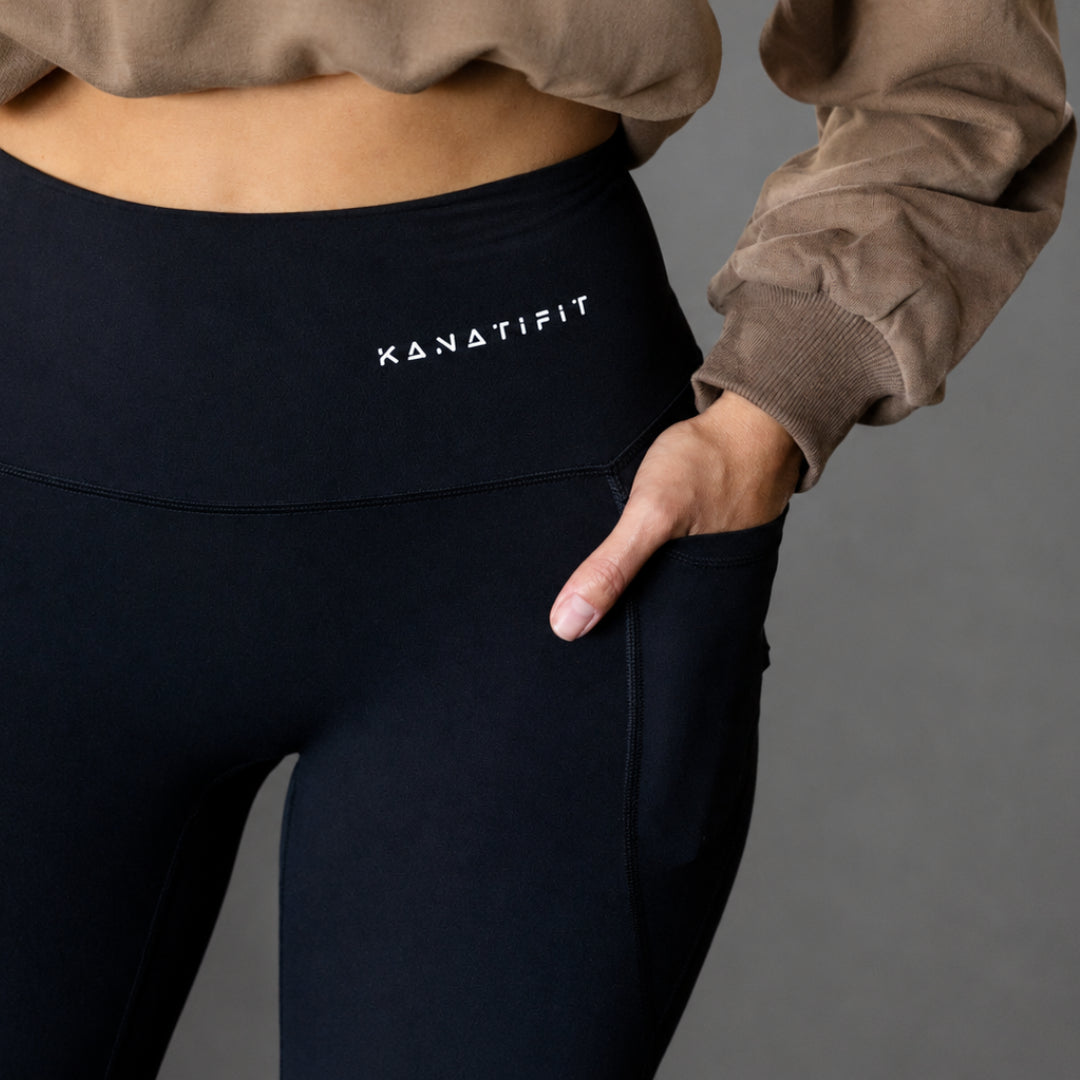 Close-up of a person wearing black high-waisted leggings with a white KANATIFIT logo on the waistband, paired with a brown sweatshirt, against a solid gray background.