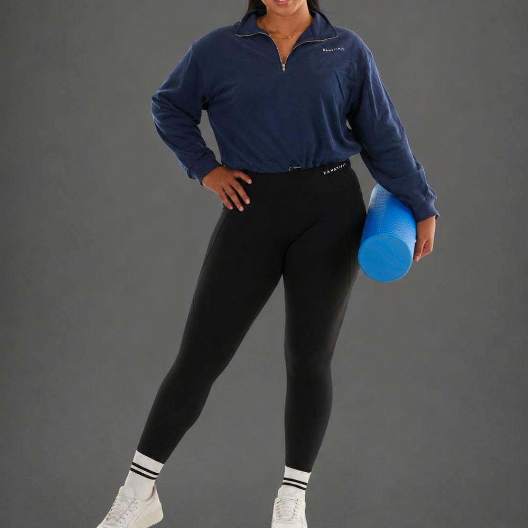 A woman modeling a navy blue quarter-zip athletic pullover and black leggings, holding a blue foam roller against a solid gray studio background.