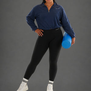 A woman modeling a navy blue quarter-zip athletic pullover and black leggings, holding a blue foam roller against a solid gray studio background.