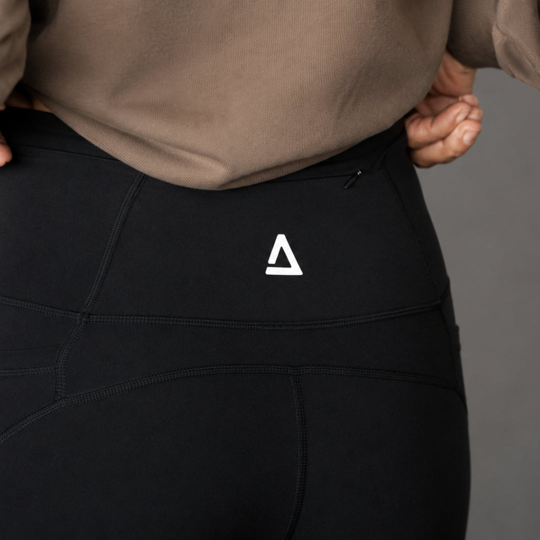 Close-up of a person wearing black high-waisted athletic leggings with a white 'KANATIFIT' logo on the back waistband, paired with a taupe sweatshirt against a neutral gray background.