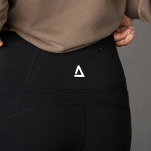 Close-up of a person wearing black high-waisted athletic leggings with a white 'KANATIFIT' logo on the back waistband, paired with a taupe sweatshirt against a neutral gray background.