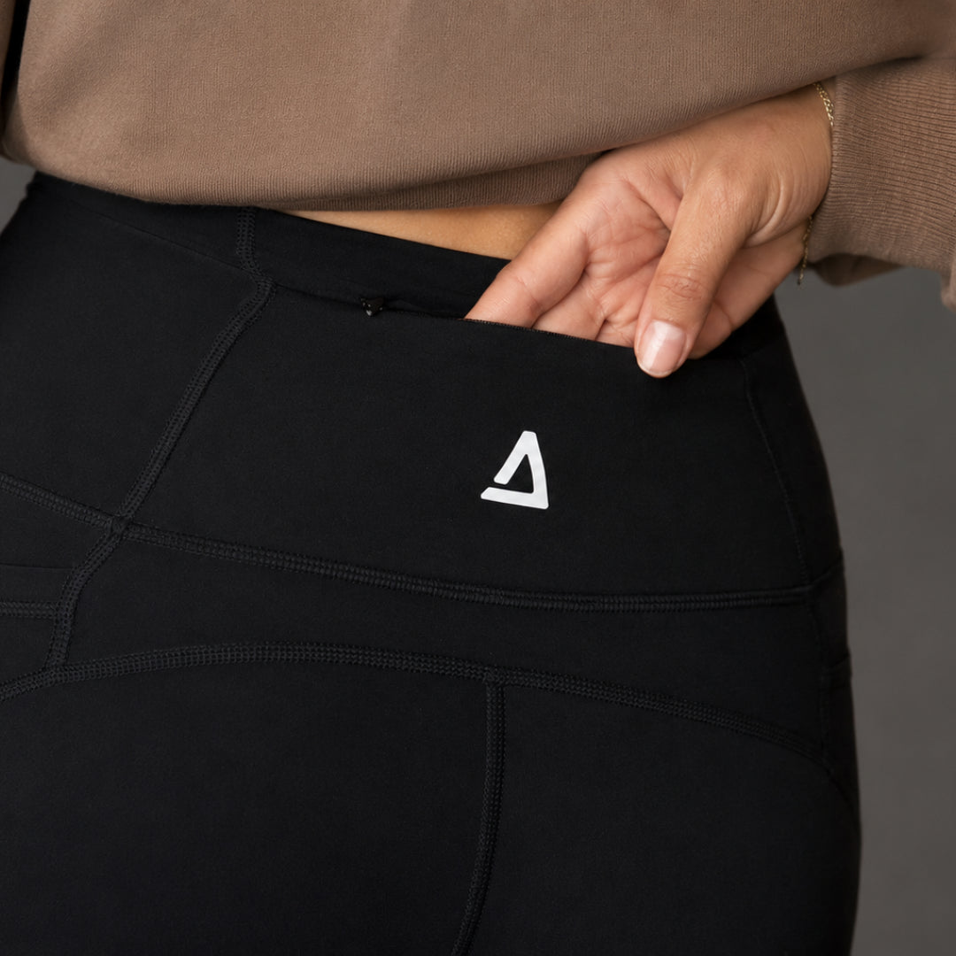 Close-up of a person wearing black high-waisted athletic leggings with a white triangular 'KANATIFIT' logo on the side pocket, shown with a hand reaching into the pocket. The person is also wearing a brown top.