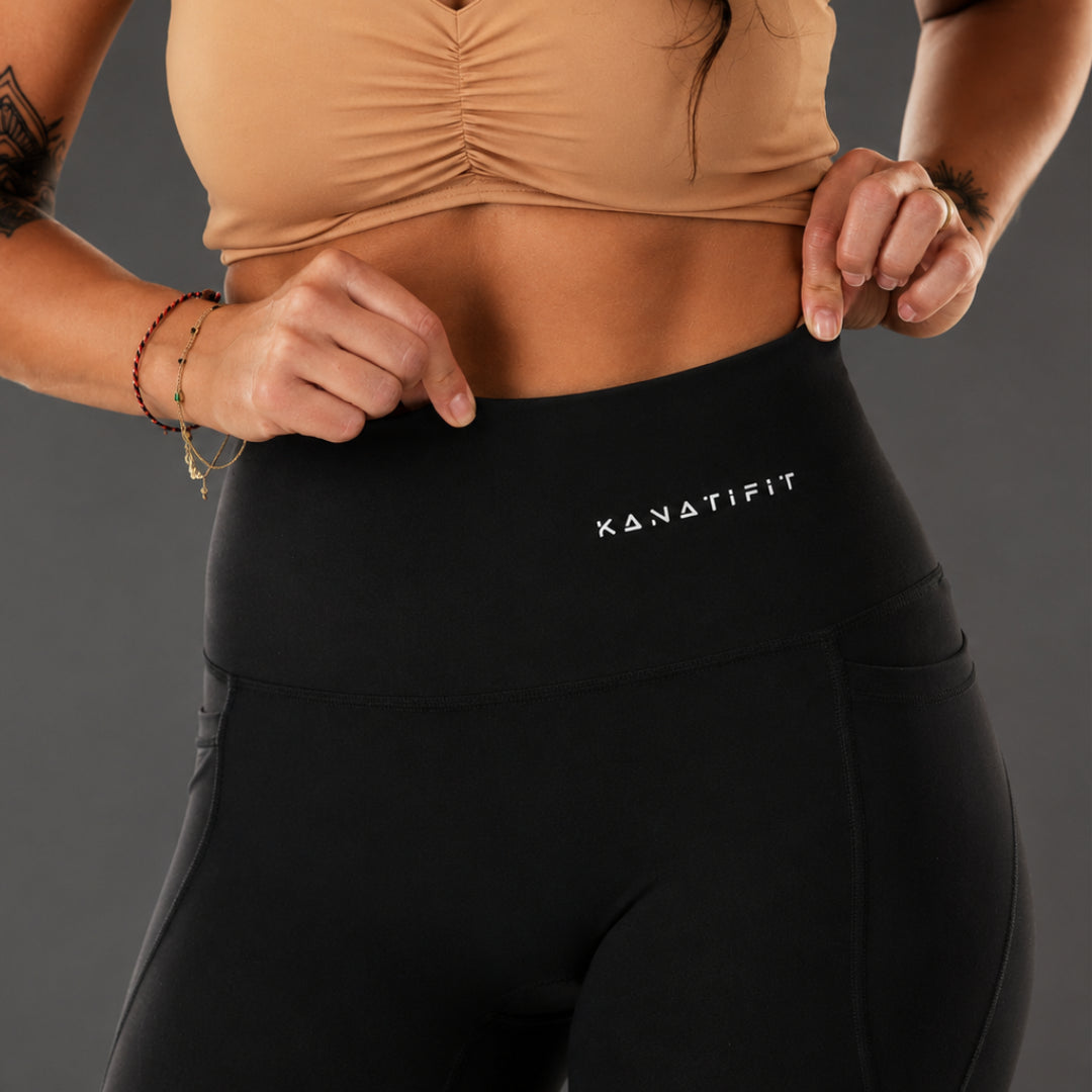 Close-up of a model wearing black high-waisted leggings with the white 'KANATIFIT' logo on the front, paired with a beige ruched sports bra against a dark gray studio background.