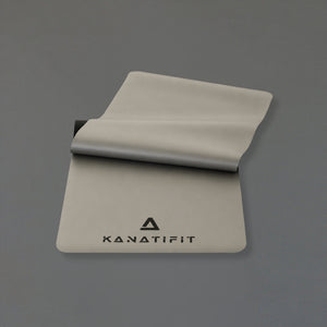 A light gray yoga mat with the KANATIFIT logo printed in black, shown partially unrolled on a solid gray background.