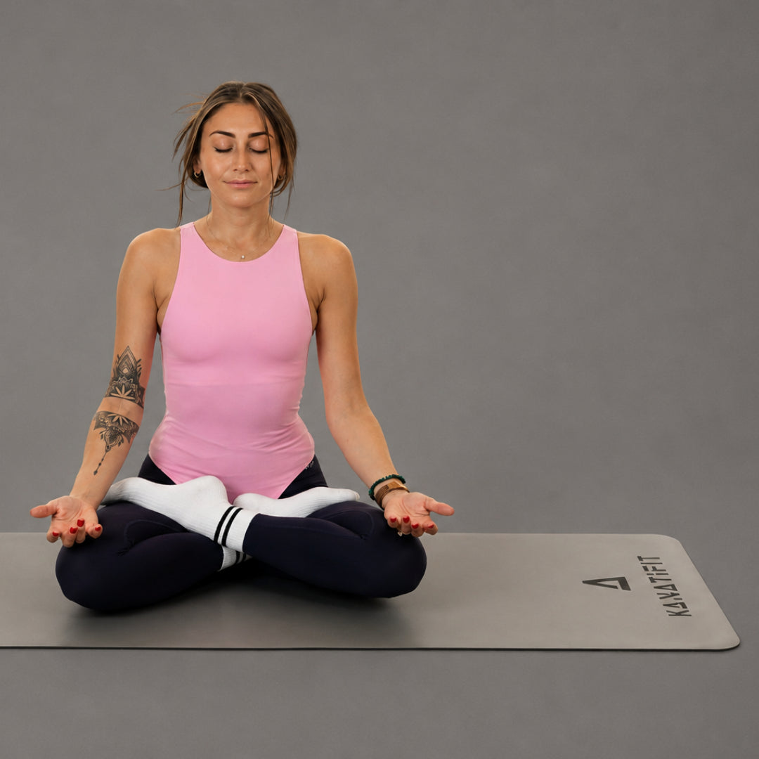 A woman in a pink tank top and black leggings sits cross-legged in a yoga pose on a gray yoga mat with the brand name 'KANATIFIT' printed on it, against a solid gray studio background.