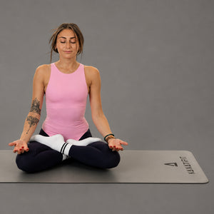A woman in a pink tank top and black leggings sits cross-legged in a yoga pose on a gray yoga mat with the brand name 'KANATIFIT' printed on it, against a solid gray studio background.