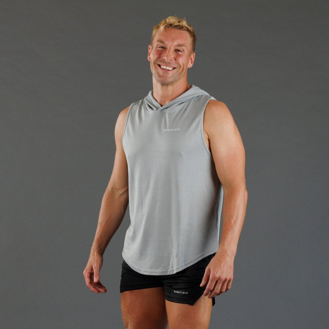 A smiling man wearing a light gray sleeveless hooded athletic shirt and black athletic shorts, standing against a solid dark gray studio background.